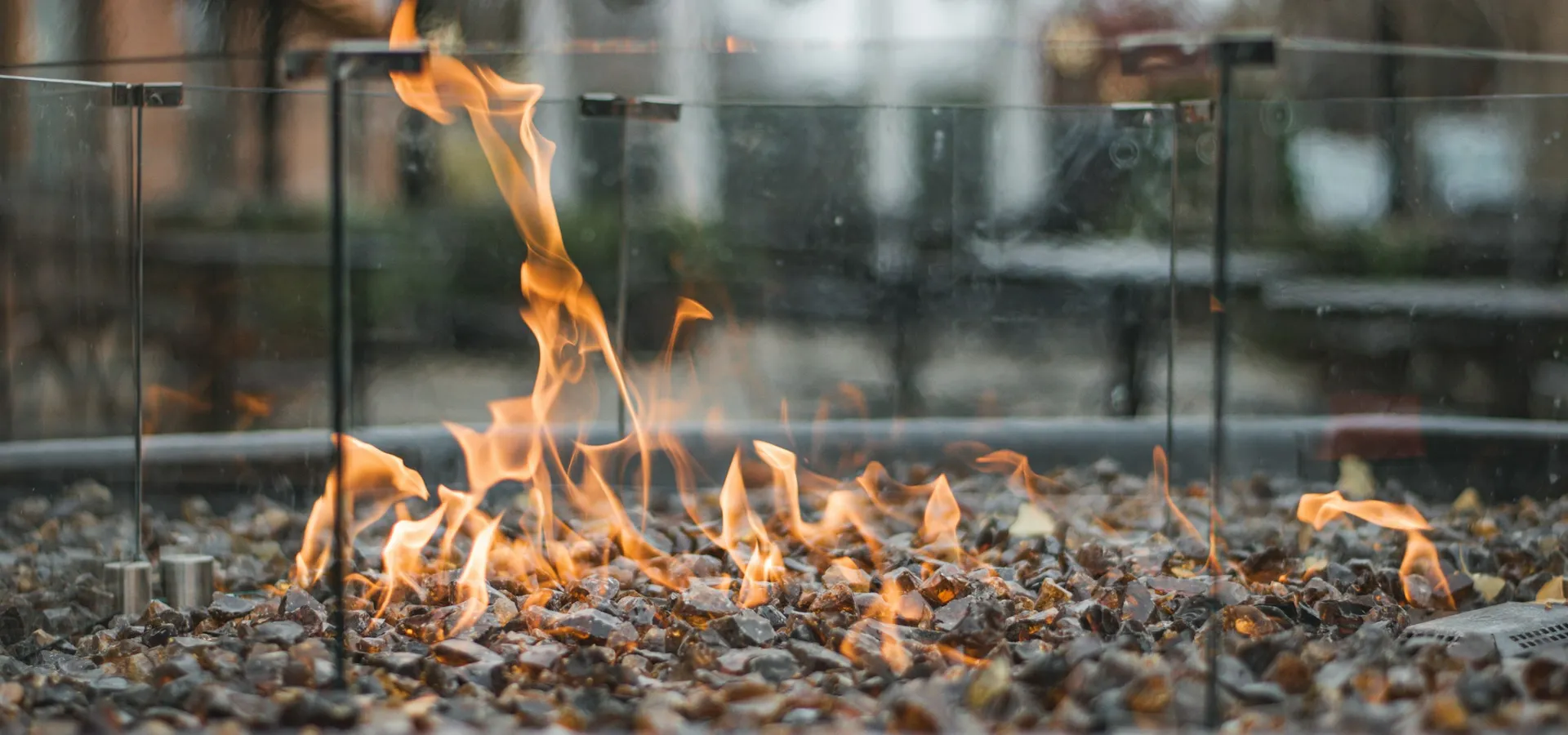 A linear gas fire pit burning across a bed of stone media, behind a glass wind guard on a terrace.