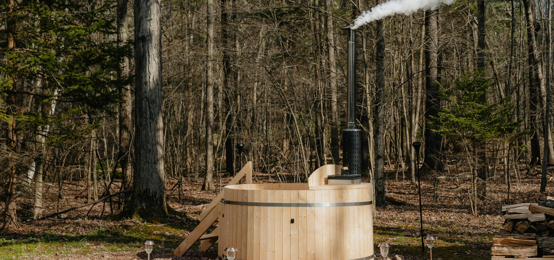 A cedar wood-fired hot tub in bare woodland, smoke rising from its flue, split logs stacked alongside.