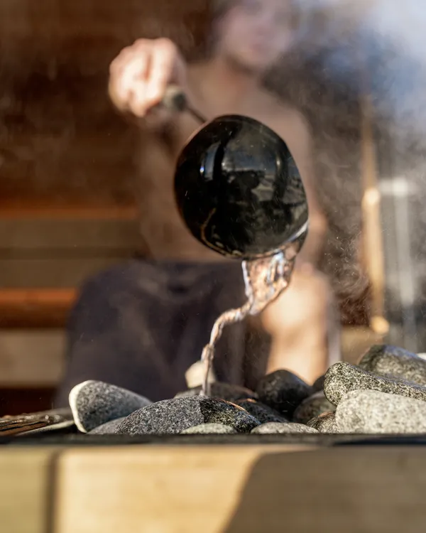 Water poured from a ladle onto the heater stones inside a cedar sauna, steam rising.