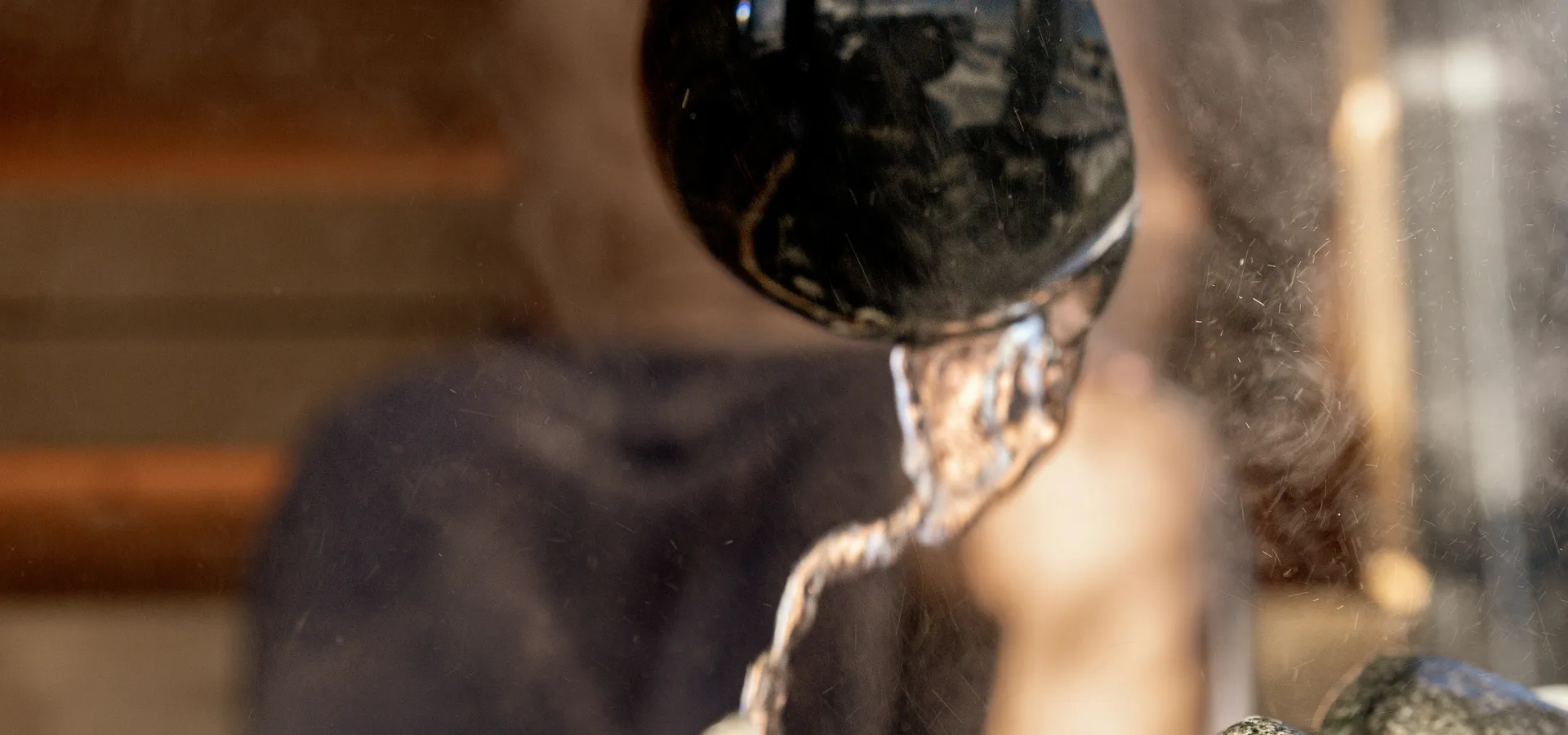 Water poured from a ladle onto the heater stones inside a cedar sauna, steam rising.