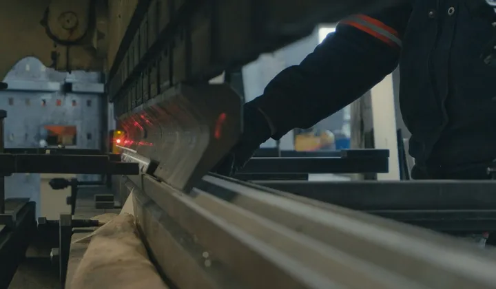 Gloved hands guiding a steel sheet through a press brake, the laser guide line visible along the bend.